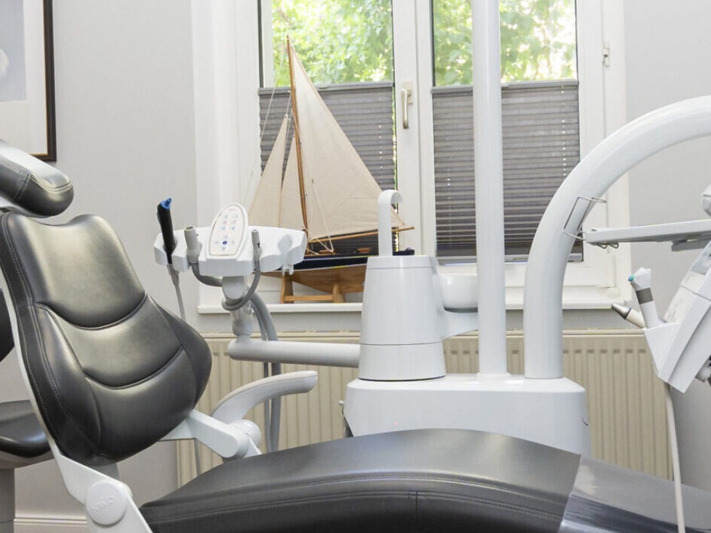 Detailansicht eines modernen Zahnarzt-Behandlungsstuhls mit Behandlungseinheit vor einem Fenster, auf dessen Fensterbank ein dekoratives Segelboot steht.