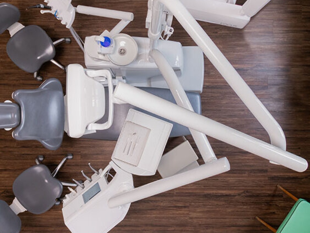 Draufsicht auf einen zahnärztlichen Behandlungsstuhl mit mehreren Schwenkarmen, Instrumenteneinheit und zwei grauen Rollhockern auf dunklem Holzfußboden.