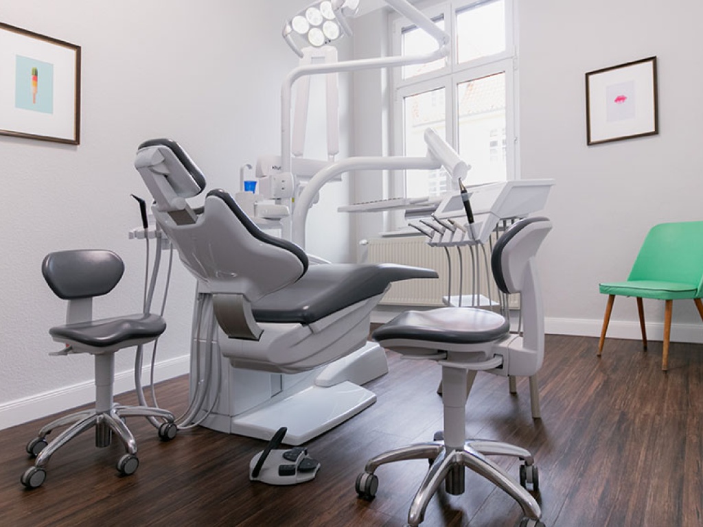 Heller, moderner Behandlungsraum einer Zahnarztpraxis mit ergonomischem Behandlungsstuhl, zwei grauen Rollhockern, Decken-Behandlungsleuchte und großem Fenster, ergänzt durch minimalistische Wandbilder und einen grünen Besucherstuhl.