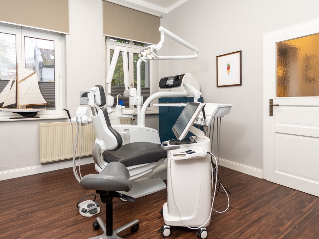 Moderner, freundlich gestalteter Behandlungsraum einer Zahnarztpraxis mit ergonomischem Behandlungsstuhl, grauem Assistenzhocker, mobiler Behandlungseinheit mit Bildschirm sowie großem Fenster und dekorativem Segelbootmodell auf der Fensterbank.