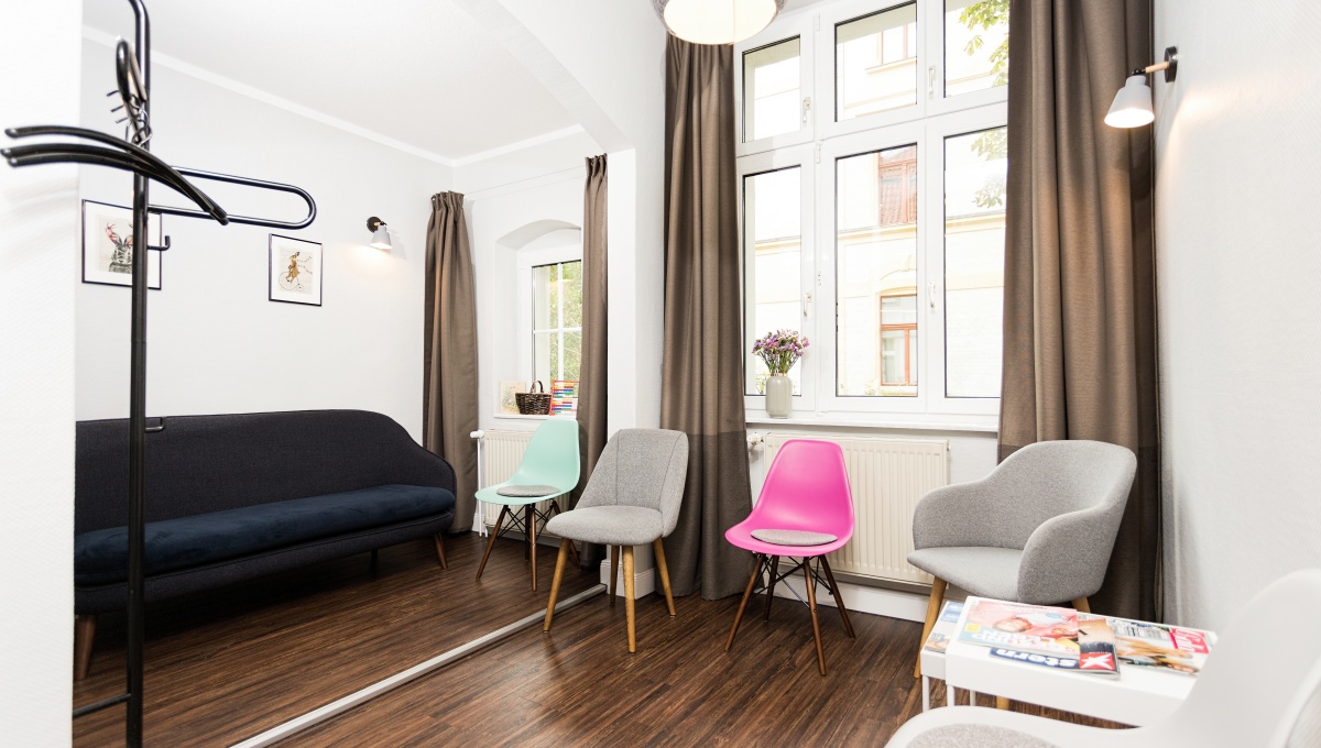 Stilvolles, helles Wartezimmer einer modernen Zahnarztpraxis mit dunkler Couch, farbenfrohen Designerstühlen, großen Fenstern, Pflanzen und angenehmer Atmosphäre für Patienten.