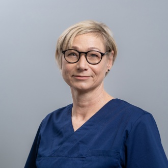 Porträt von Dentalhygienikerin Jutta Daus, erfahrene Spezialistin für professionelle Zahnreinigung und Parodontitisbehandlung, im dunkelblauen Kasack vor neutral grauem Hintergrund freundlich in die Kamera blickend