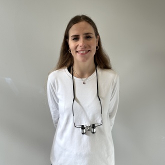 Porträt von Zahnärztin Sarah Hartmann aus DIE ZAHNARZTPRAXIS Stralsund, freundlich lächelnd im weißen Oberteil mit Lupenbrille um den Hals vor hellem Hintergrund, erfahrene Behandlerin mit Schwerpunkt Endodontie und moderner Wurzelkanalbehandlung.