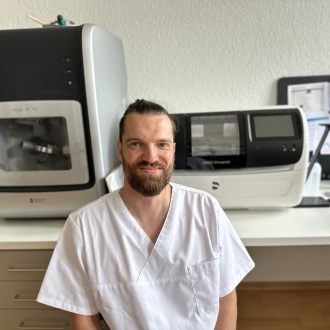 Porträt von Zahntechniker Martin Kuhnke aus dem praxiseigenen Labor der Zahnarztpraxis in Stralsund, im weißen Kasack vor moderner CEREC- und Frästechnik sitzend und freundlich in die Kamera lächelnd.