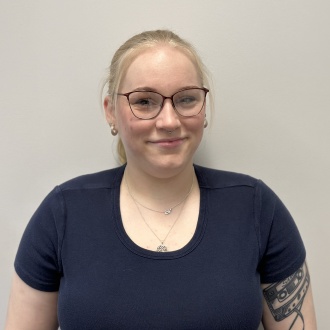 Porträt von Melissa Stresow aus DIE ZAHNARZTPRAXIS Stralsund, im dunkelblauen Oberteil mit Brille und sichtbarem Tattoo vor hellem Hintergrund, zuständig für Prophylaxe und professionelle Zahnreinigung.