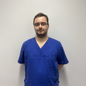 Porträt von Zahnmedizinischen Fachangestellten Tom-Manuel Garolea aus DIE ZAHNARZTPRAXIS Stralsund, im blauen Kasack vor hellem Hintergrund stehend und ruhig in die Kamera blickend.