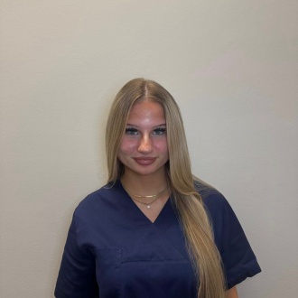 Porträt von Auszubildender Angelina Kühne aus DIE ZAHNARZTPRAXIS Stralsund, im dunkelblauen Kasack vor hellem Hintergrund, freundlich lächelnd mit langen blonden Haaren.